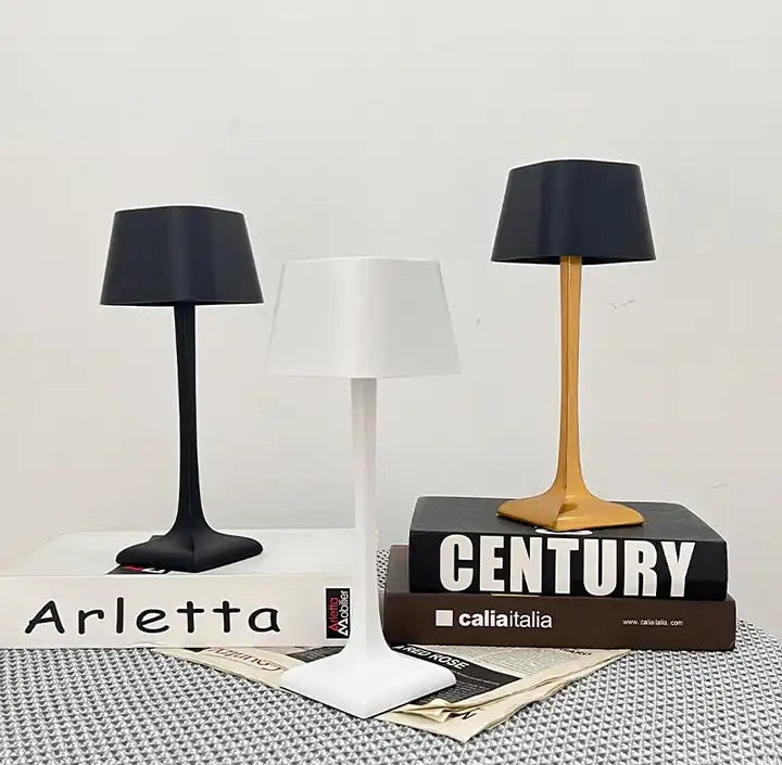 Three cordless table lamps in black white and natural wood finishes sit on a white surface next to stacked books and a sign reading Arletta