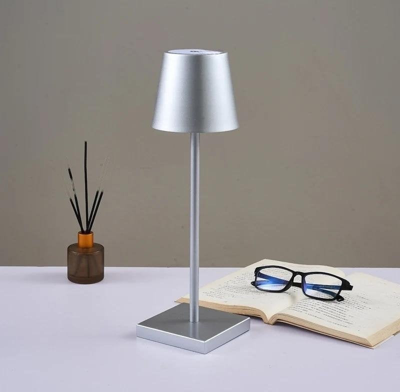 Solaris cordless table lamp in powder coated aluminium with a silver shade stands on a square base on a desk beside an open book and glasses with a small diffuser bottle in the background