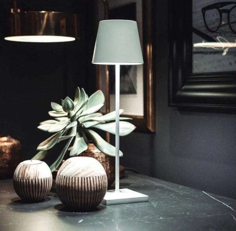 Solaris cordless table lamp in powder coated aluminium with a light grey lampshade on a white base beside a succulent plant and two round textured vases on a dark countertop