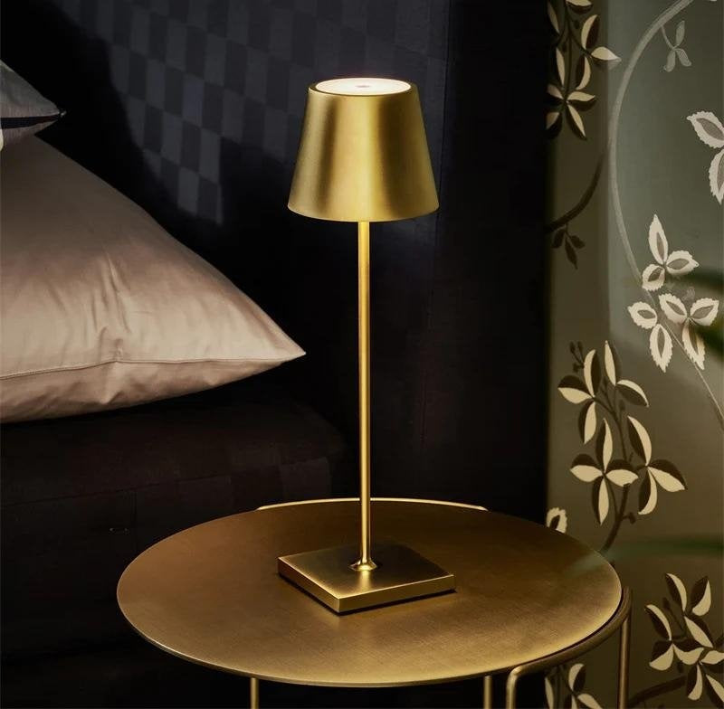 Gold cordless table lamp with a small drum shade on a slender stem and square base, placed on a round gold table in a bedroom setting