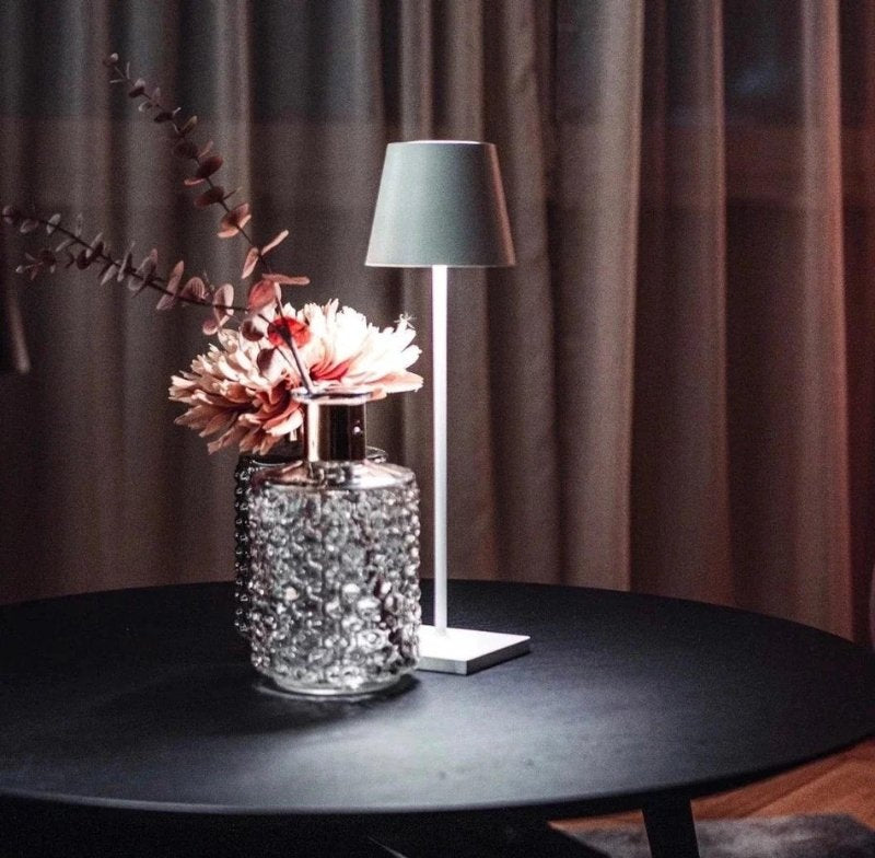 Cordless table lamp with a slim white stem and grey lampshade on a white square base sits on a dark round table beside a glass vase with crystal bead detailing and pink flowers against brown curtains.
