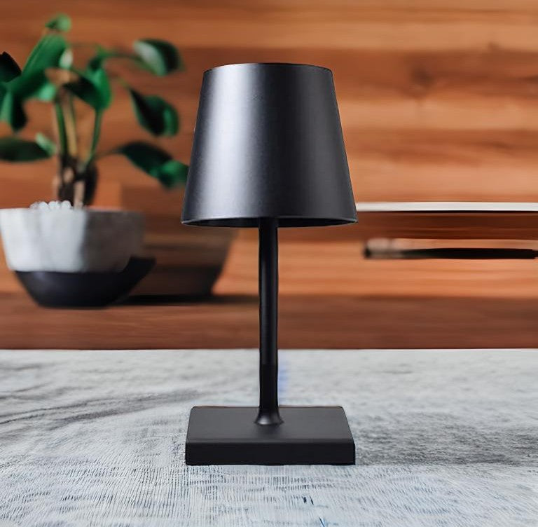 Solara touch sensor table lamp in black metal with a square base on a light desk, wood panel backdrop and a plant in the background.