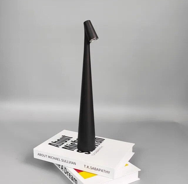 A tall black tapered wireless table lamp with a small angled head sits atop a stack of white books against a gray background.