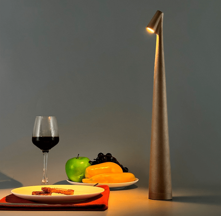Tall slender metallic table lamp with a conical base and warm light placed beside a wine glass and plates of fruit and bread on a neutral gray backdrop