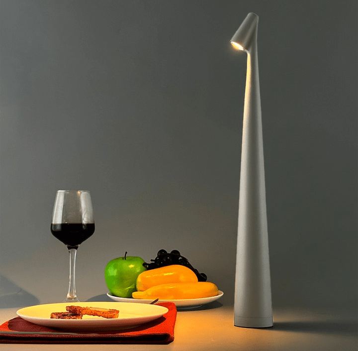Tall slender aluminum wireless touch table lamp with a dimmable downlight on a table beside a glass of red wine and a plate of fruit including a green apple and yellow peppers