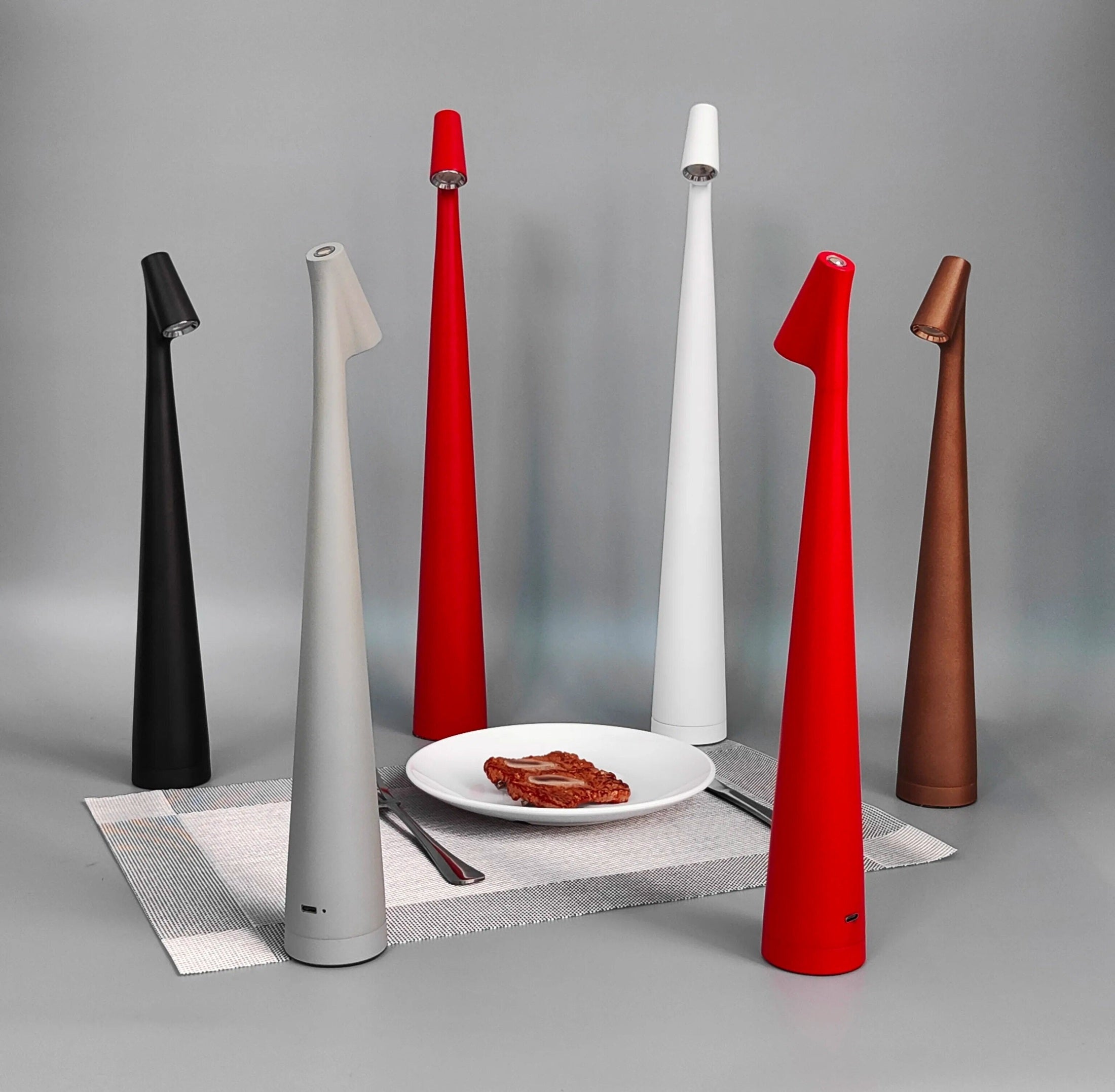 Group of tall cone shaped wireless touch control table lamps in black gray white red and brown arranged around a dining table with a plate of food and utensils on a woven placemat