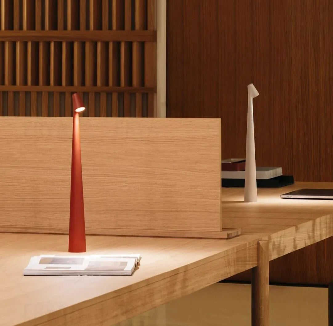 Red cone shaped wireless touch control table lamp on a light wood desk with a white tall curved lamp in the background and a magazine in the foreground.