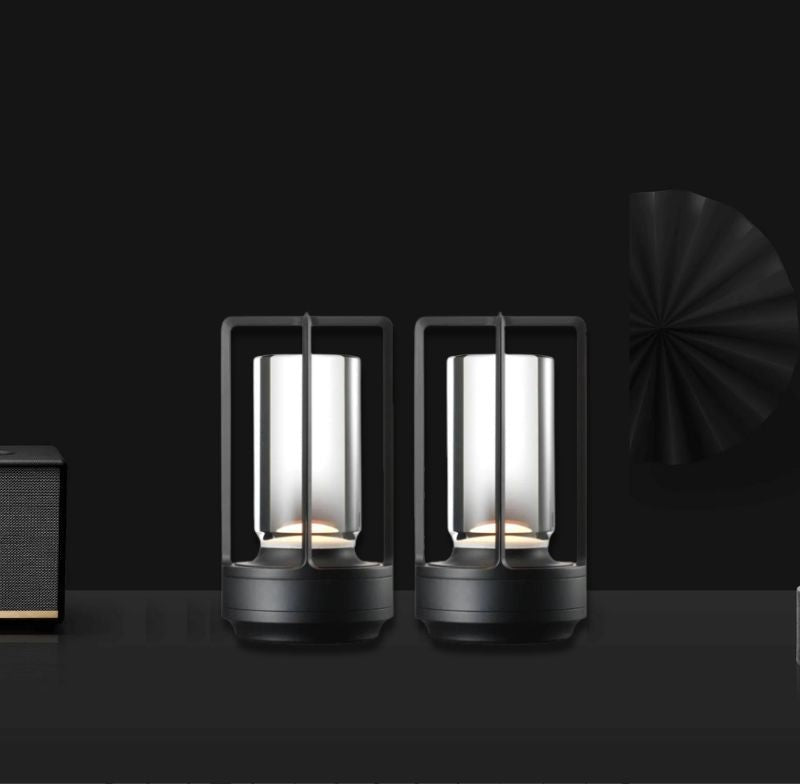Two cordless lantern lamps with metal frames and glass panels glow on a dark surface, with a speaker on the left and a black round decorative piece on the right.