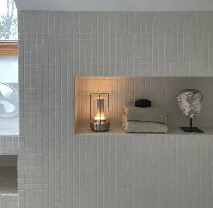 A cordless lantern table lamp with glass panels and metal frame glows warmly in a tiled wall niche beside folded towels and a decorative bust on a stand.