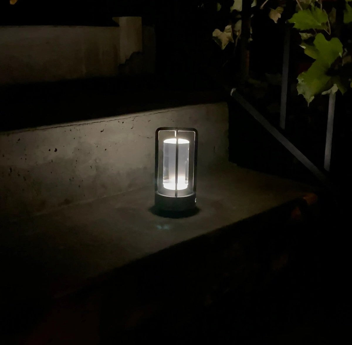 Cordless lantern with a metal frame and glass panels glowing on a concrete outdoor step at night with leaves nearby