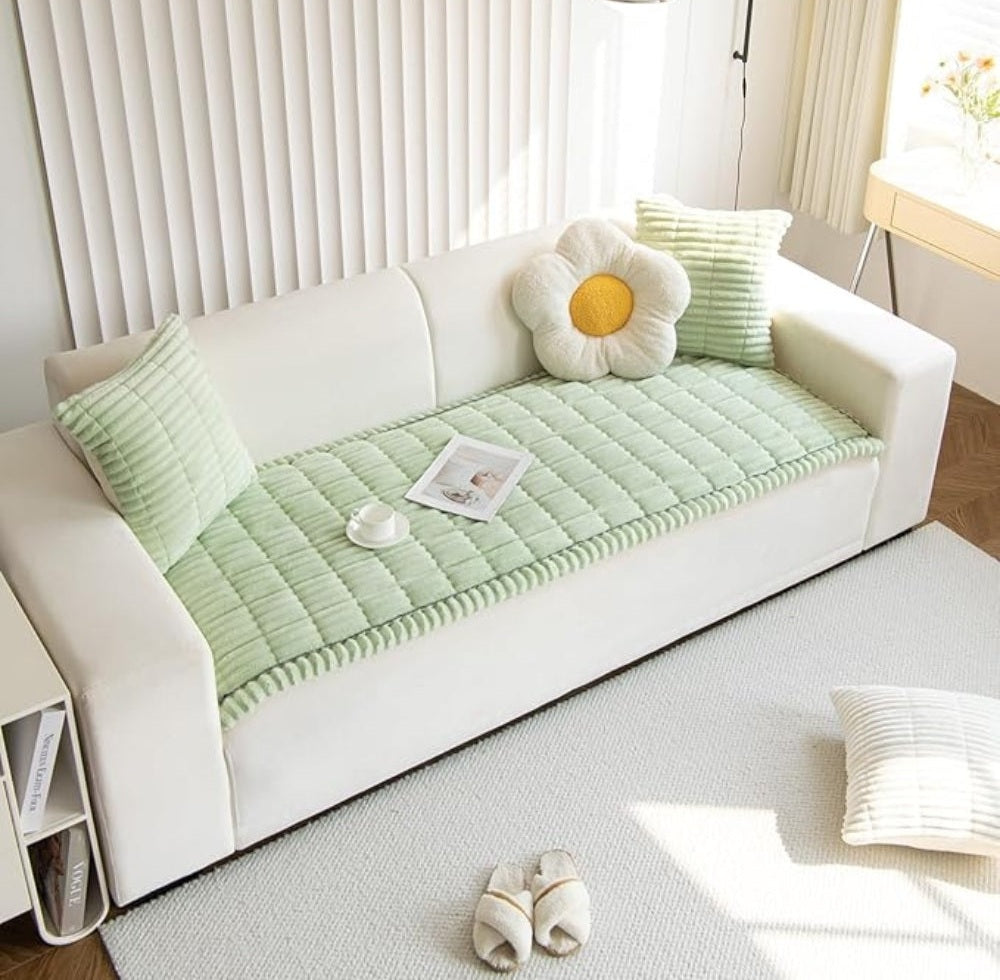 White sofa with a mint green quilted sofa cover with hand pleated edges, two green cushions, a flower shaped cushion, a magazine and cup on the cover, a side table with magazines nearby, a light gray rug, slippers on the floor, and sunlight streaming through vertical blinds.