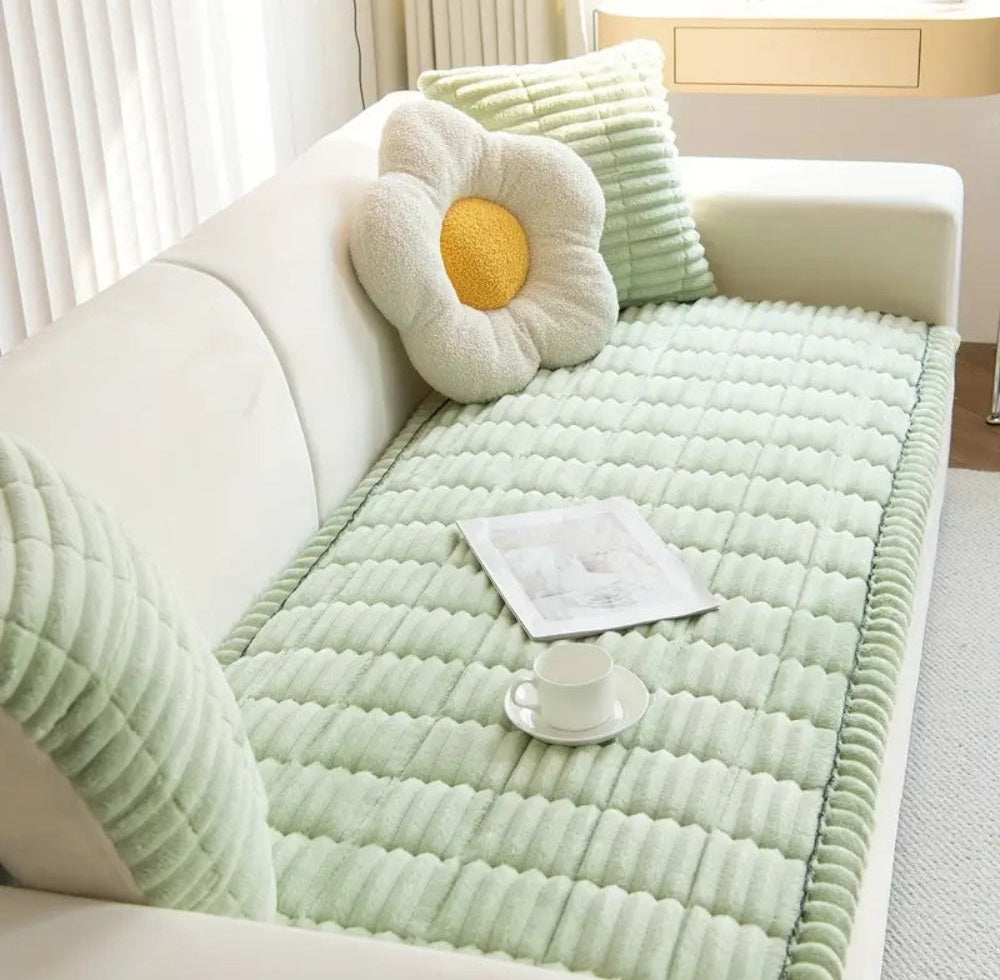 Mint green quilted sofa cover with hand pleated edges on a white sofa, paired with a cream flower shaped pillow with yellow center and a green ribbed cushion, a white cup and saucer and a magazine on top
