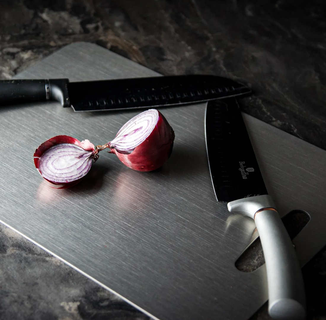 Titanium cutting board with a brushed metal surface a halved red onion and a large knife with a black blade and light gray handle resting on it