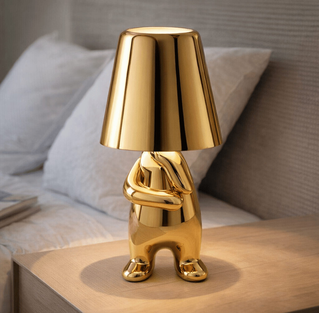Gold aluminum table lamp with a sculpted animal shaped base and a tall conical shade on a wooden bedside table in a neutral bedroom setting