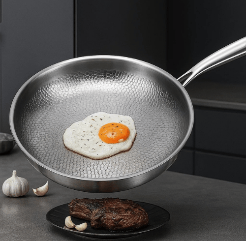 Titanium frying pan with hammered texture cooking a sunny side up egg above a plate with steak and garlic cloves