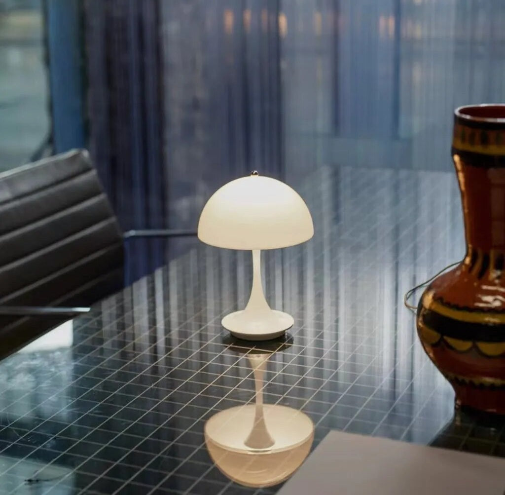 White dome shaped LED table lamp on a reflective glass table with a grid pattern, with a dark chair on the left and a ceramic vase on the right