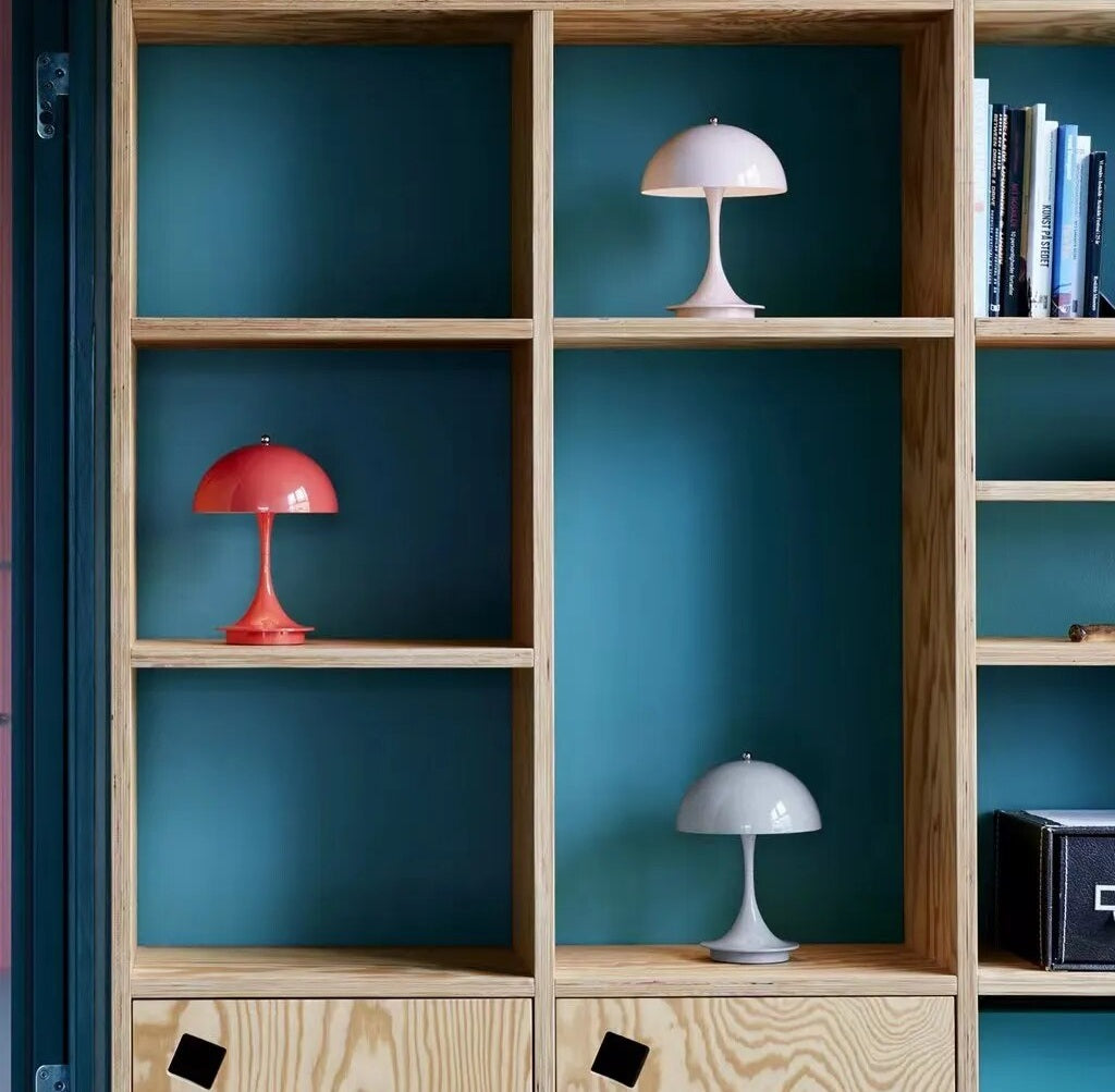 Wooden cubby shelf with teal back panels shows three dome shaped LED table lamps in red white and gray and a row of books on the right