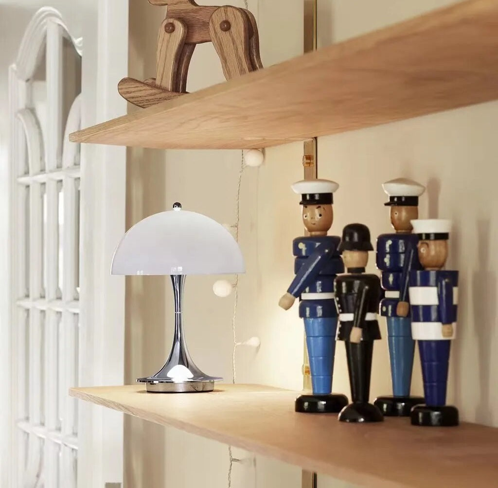 White dome LED table lamp with a chrome stem on a wooden shelf beside three wooden sailor figurines and string lights