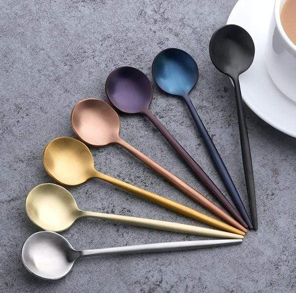 Seven plated stainless steel teaspoons in assorted colors fanned out on a gray textured surface with a white cup and saucer nearby.