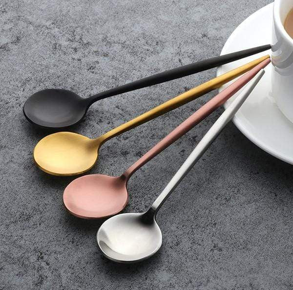 Four satin finish plated stainless steel teaspoons in black, gold, rose gold, and silver arranged diagonally on a gray textured surface beside a white cup and saucer.