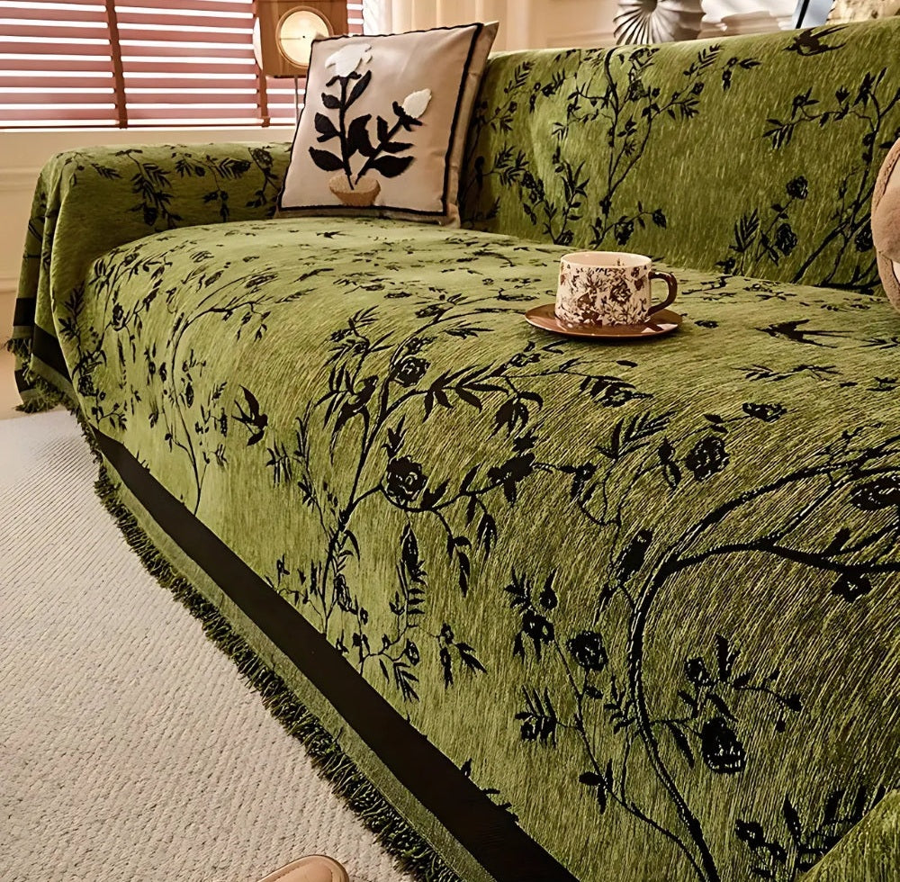 Green chenille sofa cover with black jacquard floral pattern and tassel trim on a sofa, with a decorative pillow and a teacup on a saucer placed on the cover