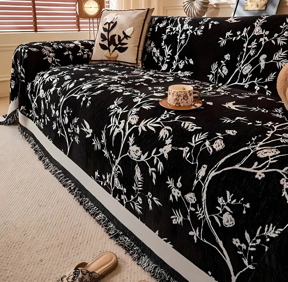 Black chenille sofa cover with white floral jacquard design and tassel trim draped over a sofa with a beige botanical cushion and a cup on a saucer resting on the cover.