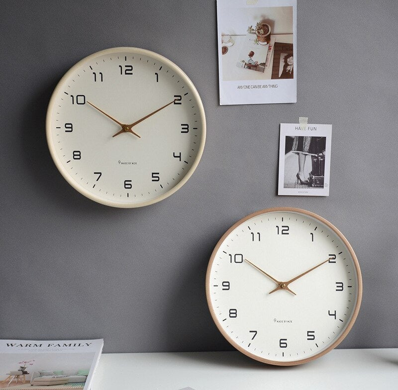 Two Nordic minimalist wall clocks with white faces, black numerals and gold hands, a cream frame clock on the left and a light wood frame clock on the right, mounted on a gray wall with photos attached beside them