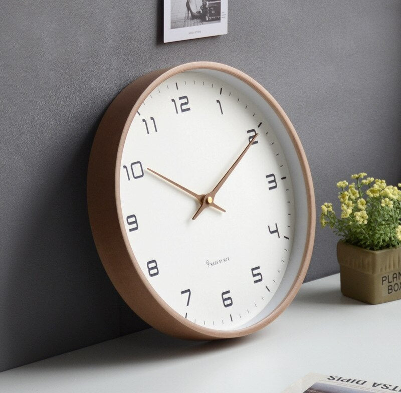 Nordic minimalist round wall clock with a white dial and copper hands, light wood frame on a white surface against a gray wall with a small potted plant and a framed photo nearby