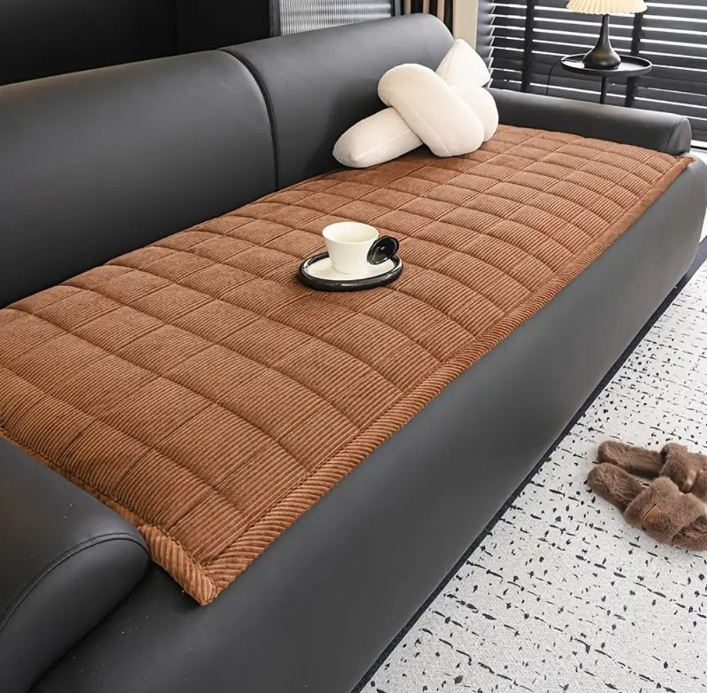 Brown quilted sofa cover mat on a dark gray leather sofa with white pillows and a cup on a saucer.