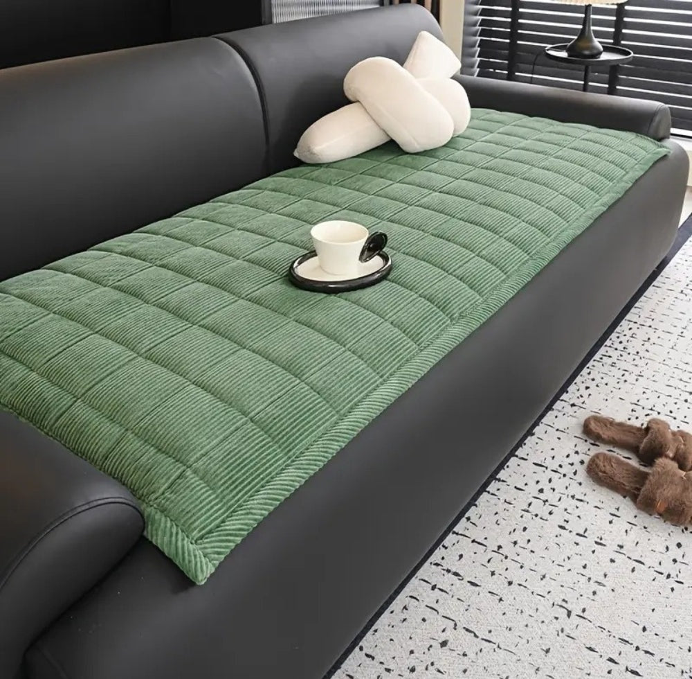 Green quilted sofa cover mat with non slip backing on a black leather sofa, white cushions, a white cup on a saucer, and brown fluffy slippers on a light speckled rug