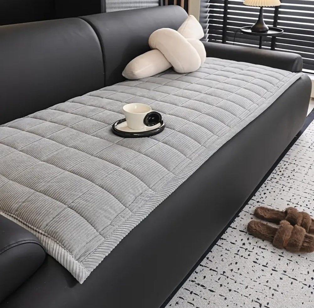 Gray quilted microfiber sofa cover mat on a black leather sofa with a white cup on a tray resting on the mat and beige cushions in the background