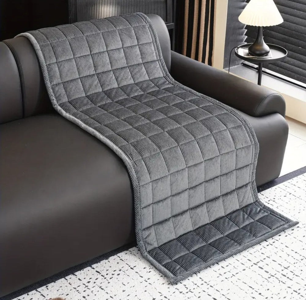 Gray quilted sofa cover mat with a non slip backing draped over a dark leather sofa featuring a square quilted pattern