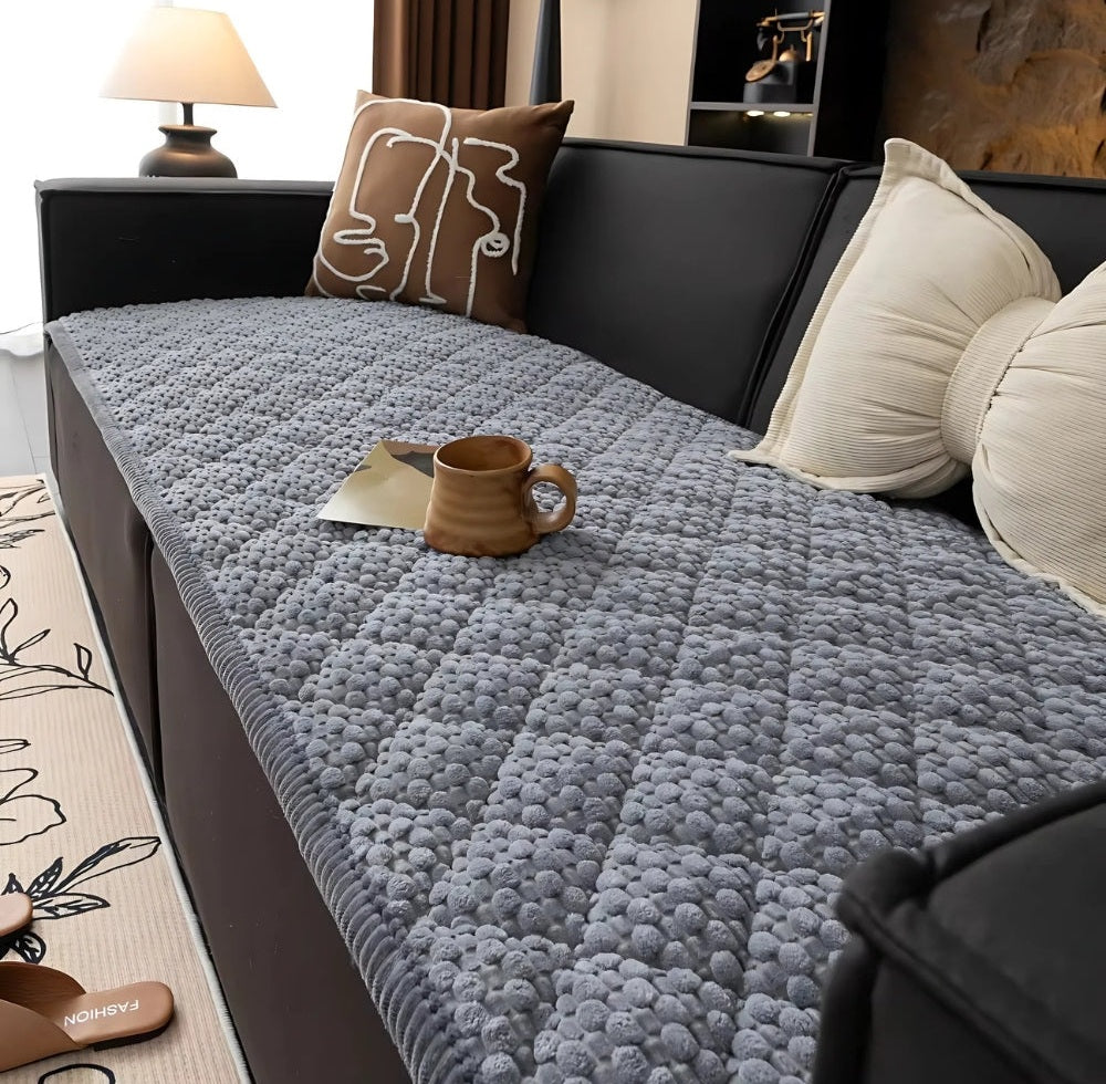 Grey bubble textured sofa protector slipcover on a dark sofa with cream and brown pillows, a ceramic mug on a small card on the cover, and a lamp and rug in the background.