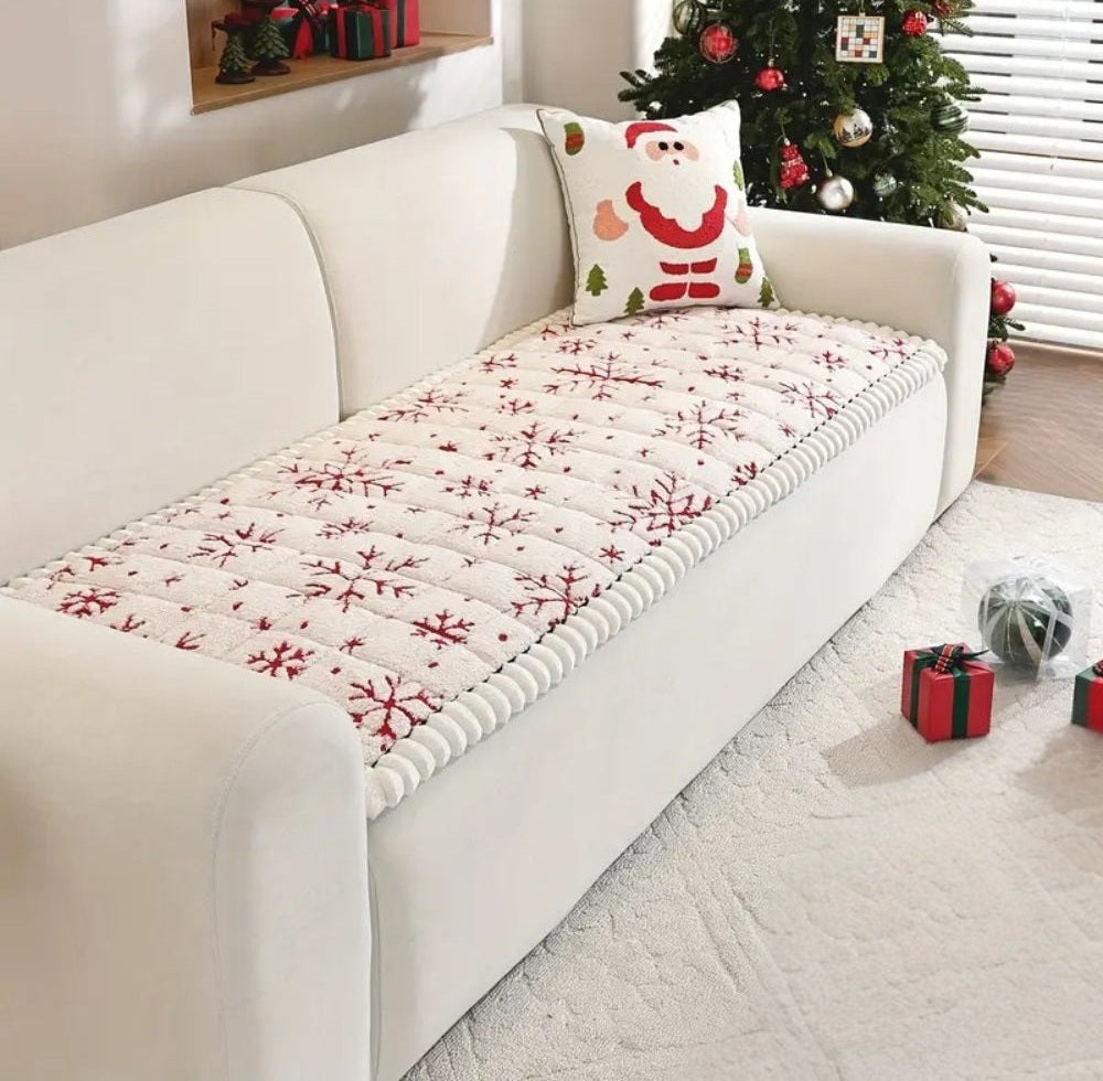 White sofa with a red snowflake quilted cover and a Santa pillow, a Christmas tree and wrapped gifts nearby.