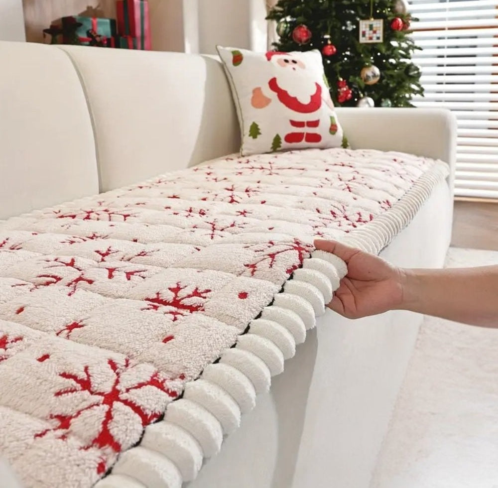 Cream quilted sofa cover with red snowflake pattern on a white sofa; a hand pulls the thick edge while a Santa pillow and a Christmas tree appear in the background