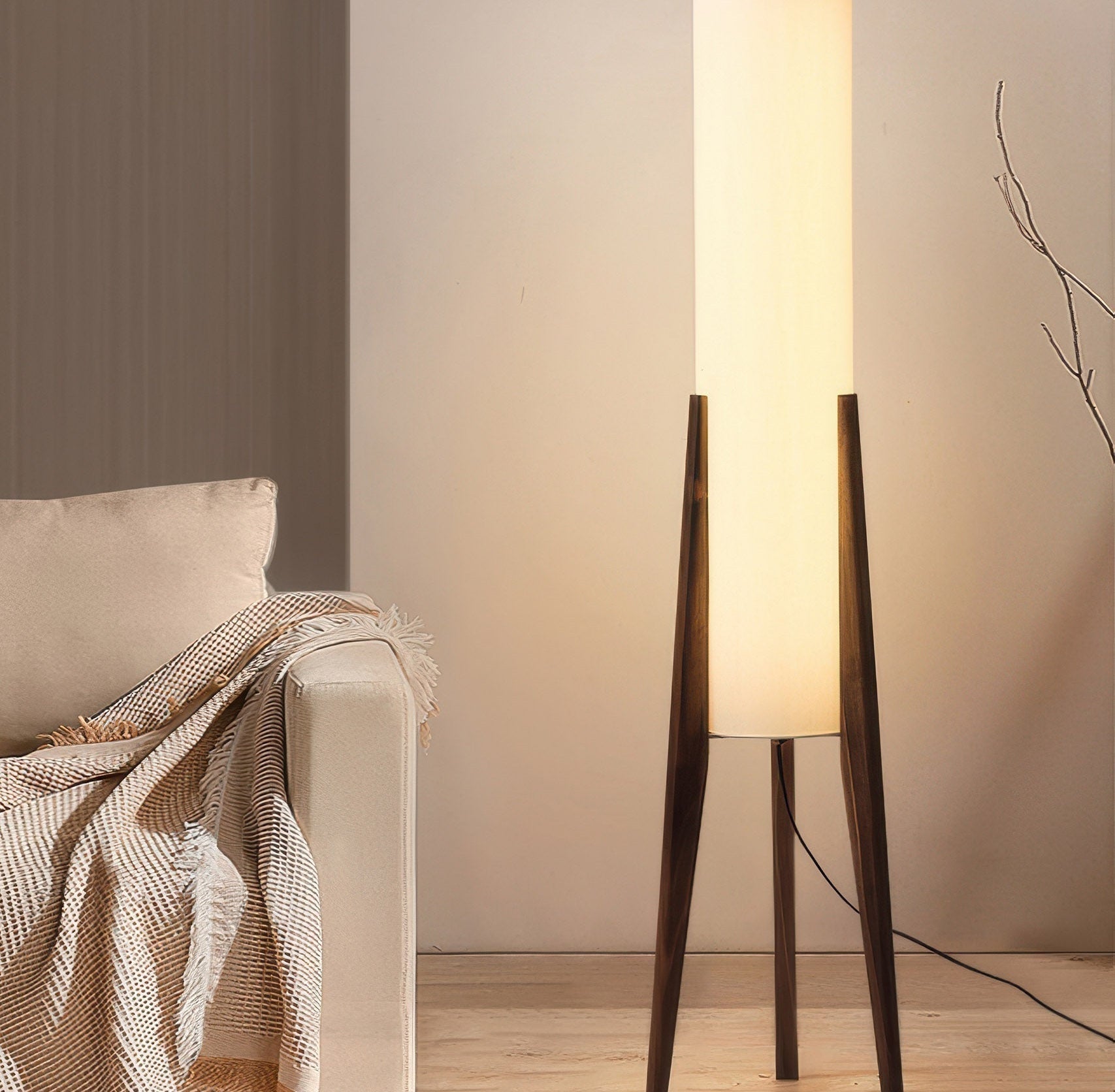 A tall Japandi floor lamp with a cream fabric shade and dark walnut legs standing beside a beige sofa with a fringed woven throw blanket