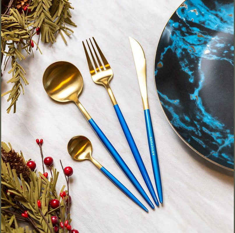 Blue handle gold plated cutlery set arranged on a white surface beside a blue marbled plate with greenery and red berries