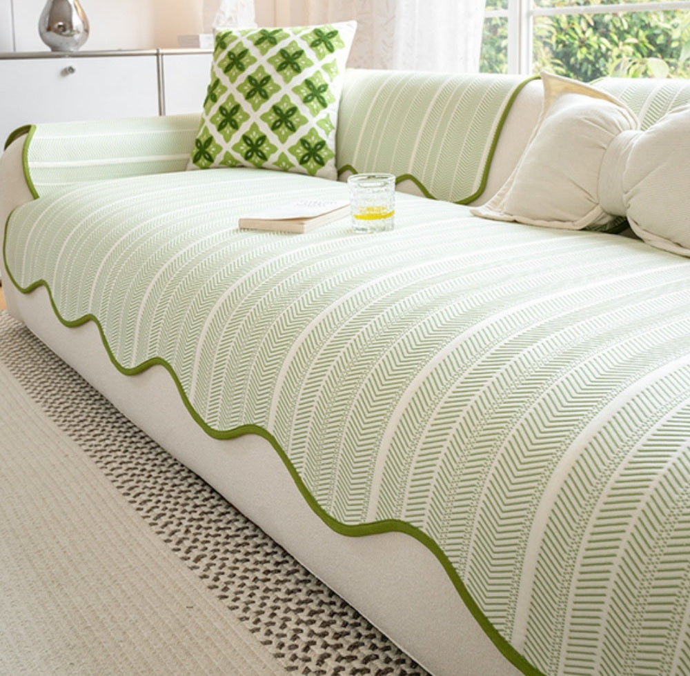 Light green herringbone microfiber sofa cover with a scalloped green wave edge trim on a white sofa, beige cushions a green patterned throw pillow and a book with a glass of lemon water on the seat