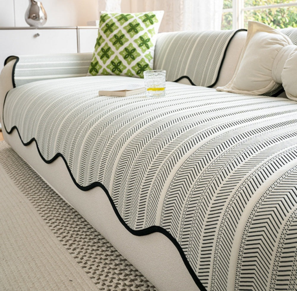 White sofa draped with a Marais herringbone microfiber blend sofa cover with scalloped wave edge trim, a green patterned cushion, and a glass with a lemon slice beside a book