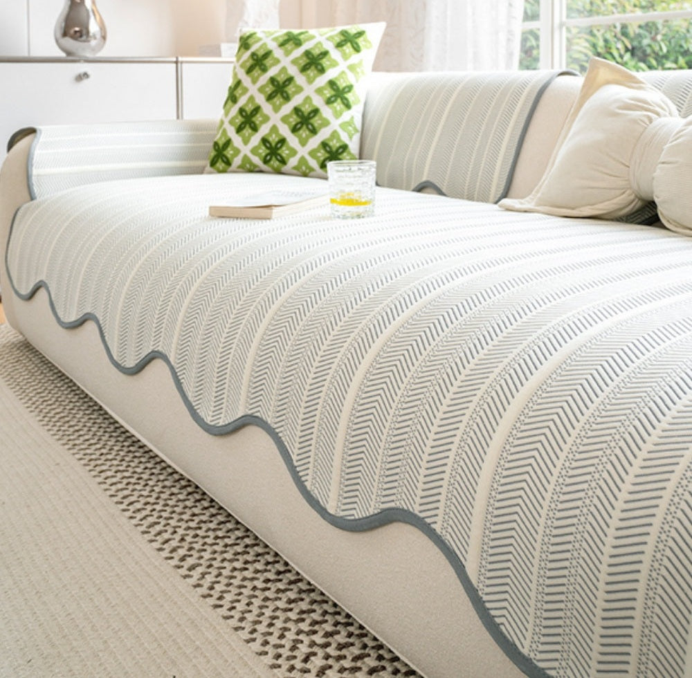 Sofa covered with a white microfiber blend fabric in a herringbone pattern and blue scalloped wave edge trim, with a green patterned pillow and a small book and glass on the arm.