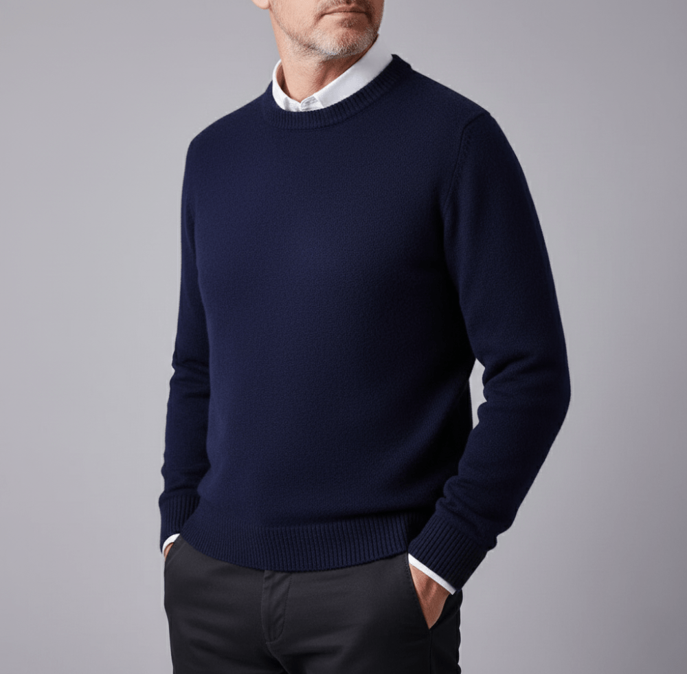 Man wearing a navy blue cashmere pullover sweater with a classic crewneck, white shirt collar peeking out, and dark trousers against a gray background