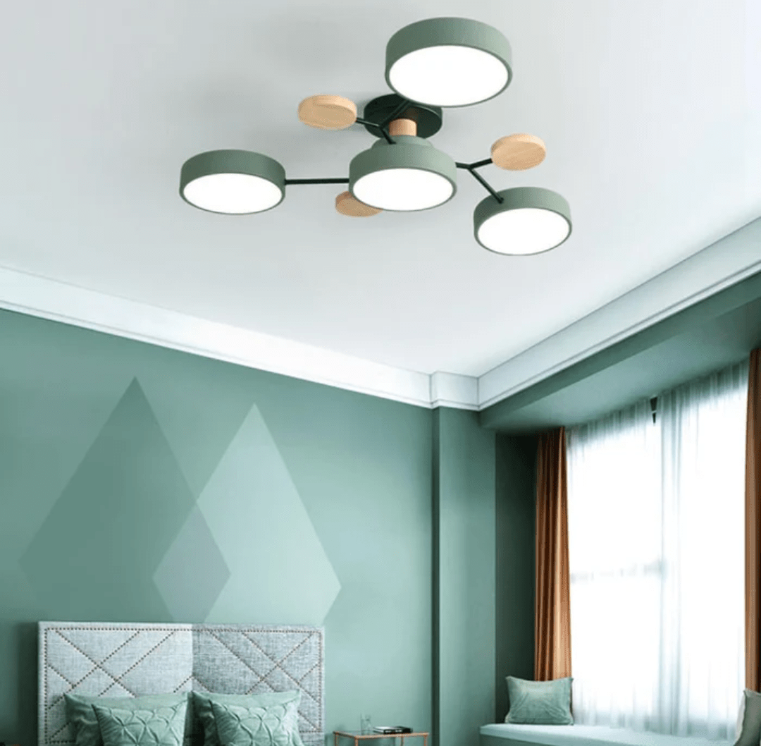 A mint green bedroom with a five light round LED ceiling fixture in green with wooden accents, a quilted bed headboard with green pillows, and a window with white sheer curtains and brown drapes