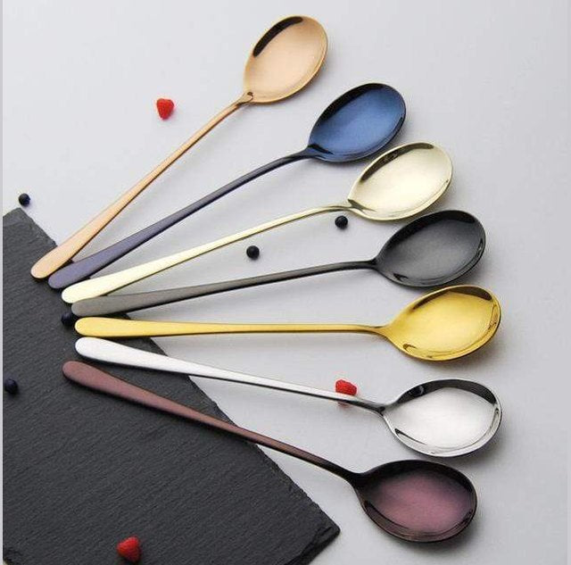An assortment of stainless steel spoons with gold silver rose gold blue black and purple plated finishes arranged in a curved fan on a light background beside a dark slate corner