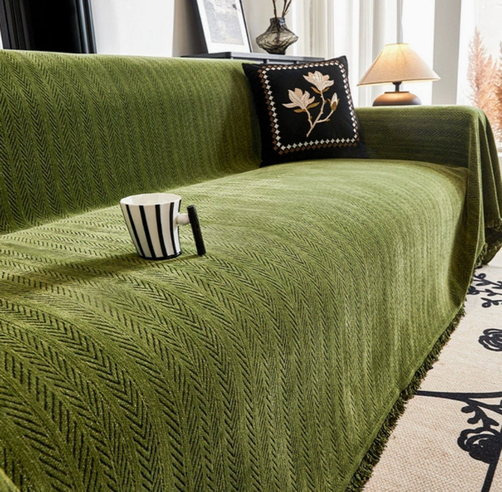 Green herringbone sofa cover with tassel trim on a sofa, a decorative black pillow with floral embroidery, a striped mug resting on the cover, and a table lamp in a sunlit living room