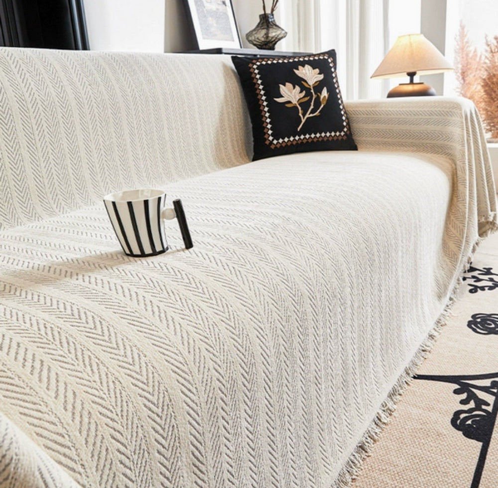 Cream sofa covered with a herringbone sofa cover featuring tassel trim and a black decorative pillow with gold floral embroidery, a striped mug on the cover, a side table lamp, and a patterned rug nearby.