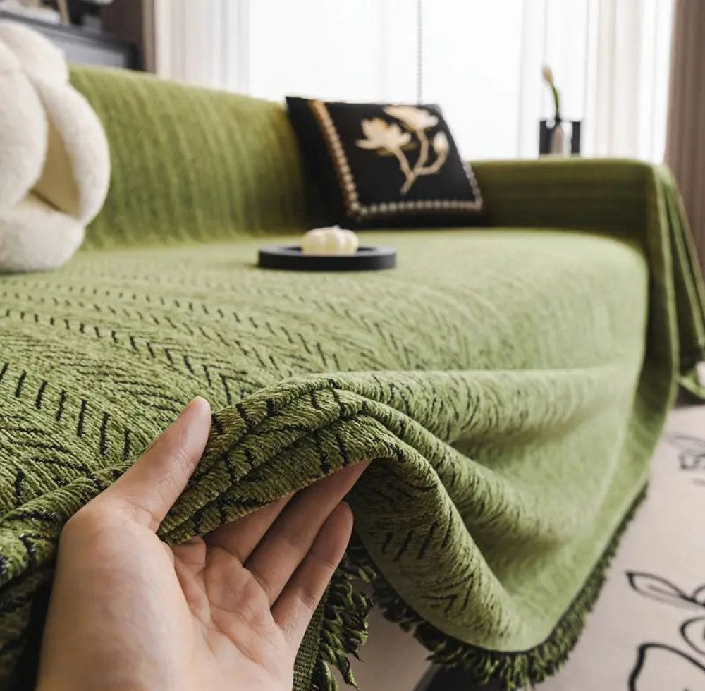 Sableweave Herringbone Sofa Cover in green microfiber blend chenille with tassel trim draped over a sofa with a hand lifting the edge to reveal the fringe while a black decorative pillow and a rolled white blanket rest on the couch