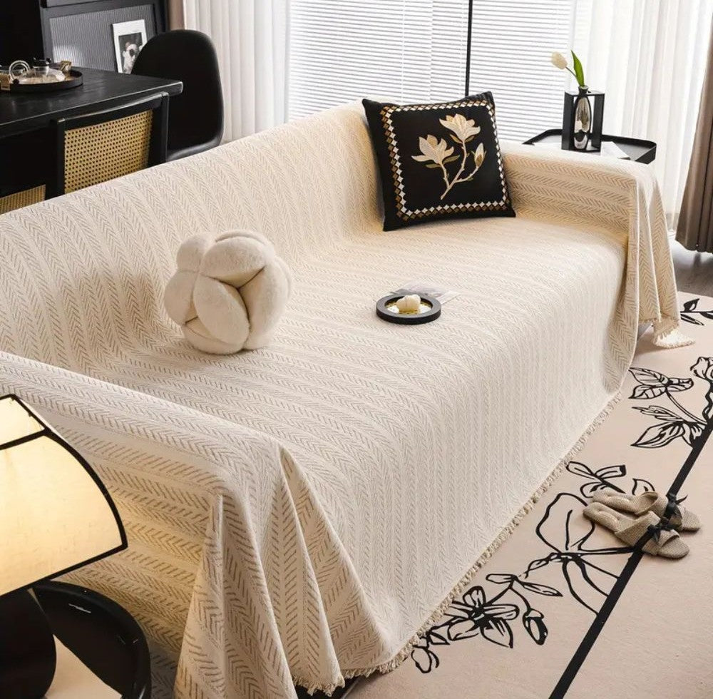 Cream herringbone sofa cover with tassel trim on a sofa, a knotted round cushion, and a black floral embroidered pillow in a bright living room with a patterned rug.