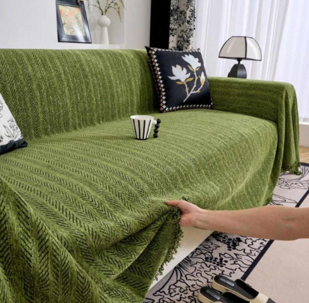 Green herringbone sofa cover with tassel trim draped over a sofa, black pillow with white floral design on the back, striped cup on the cover, and a hand pulling the corner to adjust
