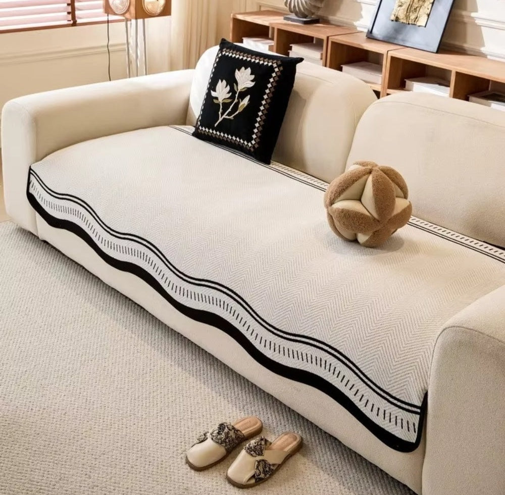 Beige sofa with a light microfiber cover featuring a black wavy edge trim, a black cushion with white leaf pattern, a knotted beige pillow, and beige slippers on a light carpet.