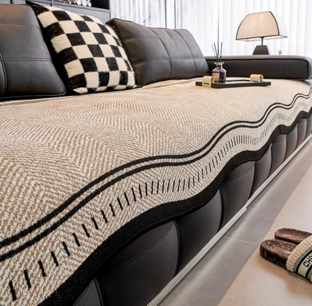 Beige woven sofa cover with a black wavy border on a black leather sofa, paired with a black and white checkered pillow and a small tray on the armrest